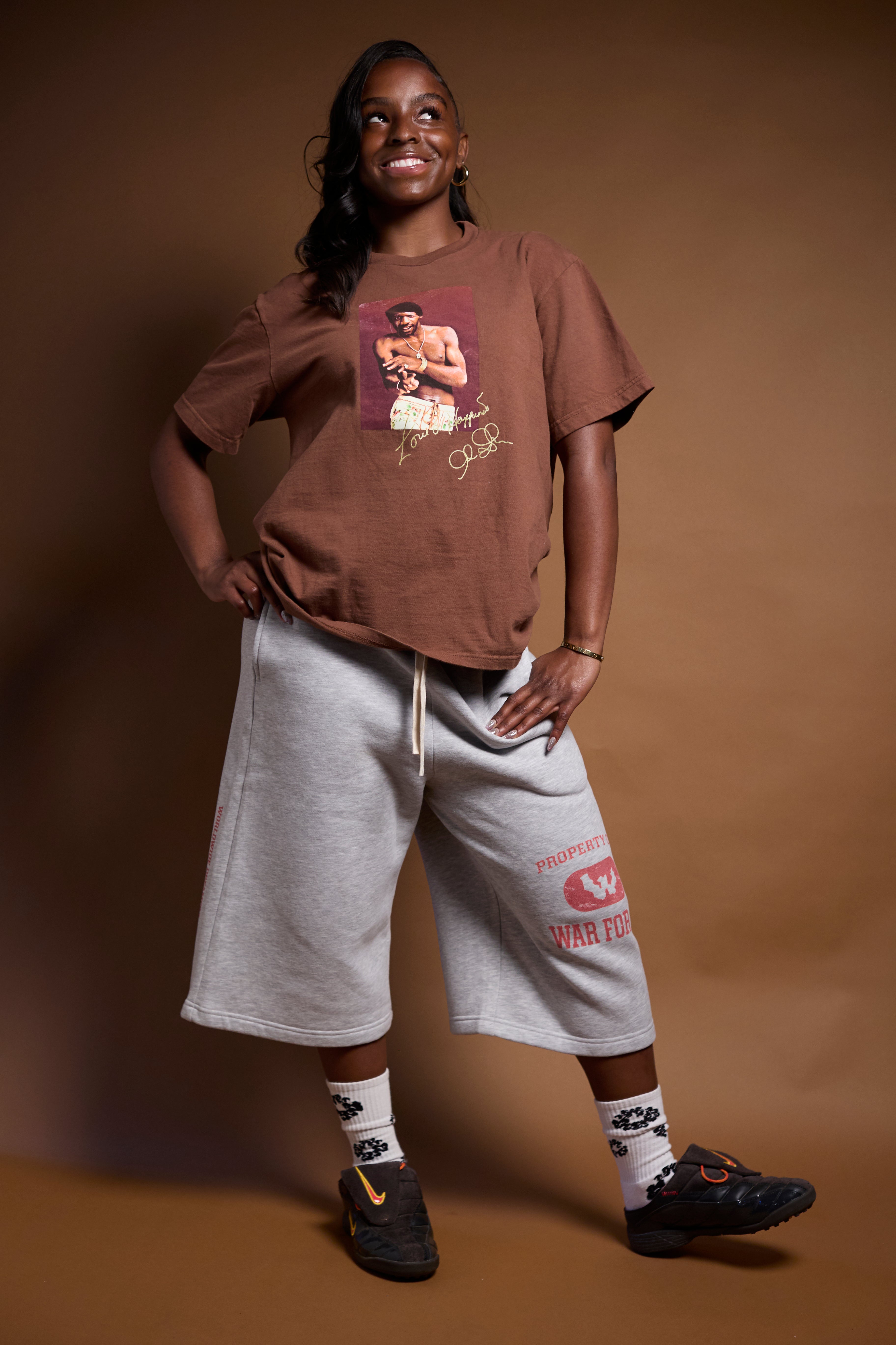 Person wearing a brown t-shirt with a graphic design and gray shorts on a brown background